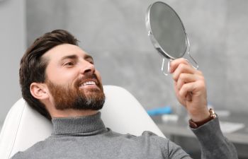 Happy man in a dental chair looking at his perfect smile achieved with dental implants in a mirror.