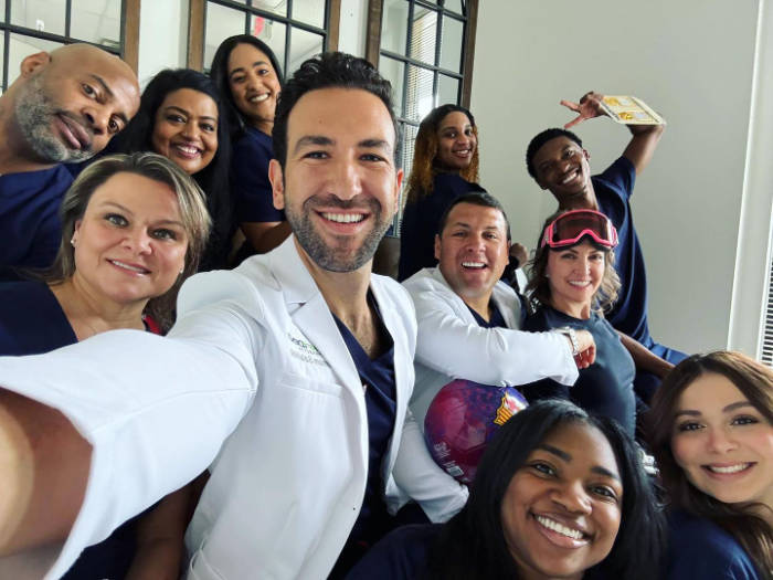 Group selfie of diverse healthcare workers or medical staff in scrubs and white coats, smiling and posing together in what appears to be a medical facility with large windows.
