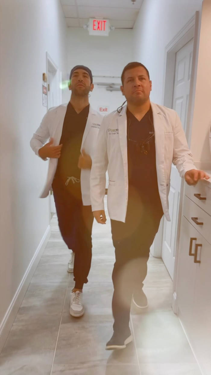 Two men in white medical coats walking down a hospital hallway with exit sign visible overhead.