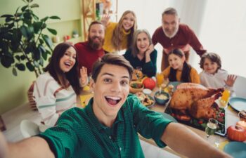 A group of people sit around a table with a roast turkey, smiling and waving at the camera for a selfie in a bright dining room.