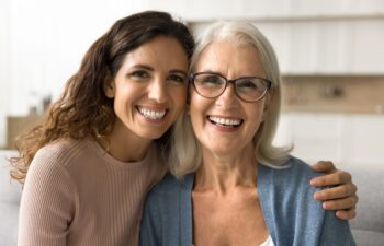 Cheerful beautiful adult daughter woman hugging happy blonde senior mom. Both are looking at camera with perfect toothy smile.