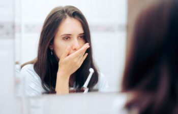 A concerned woman with toothbrush checking her breath in front of a bathroom mirror.